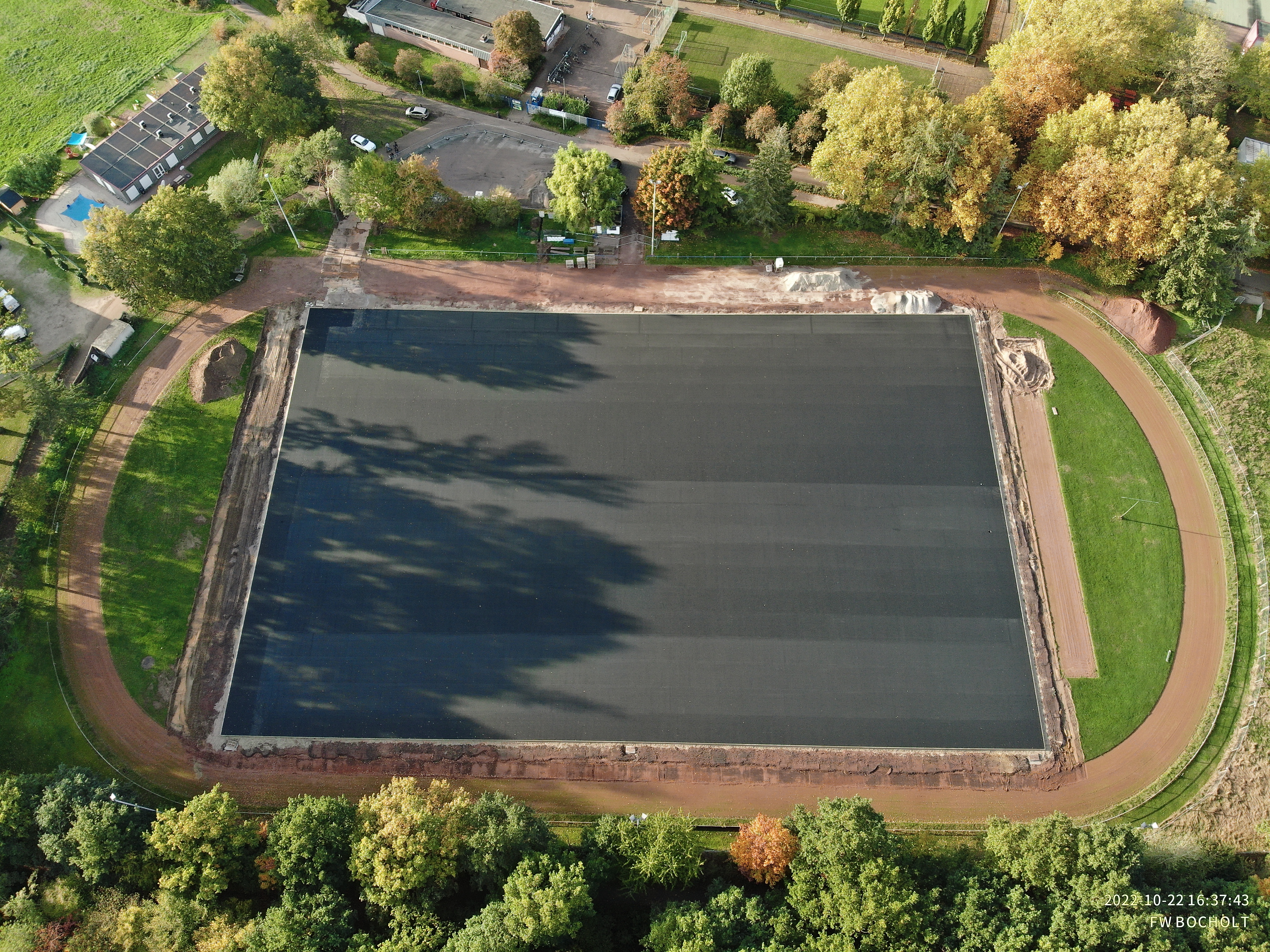 Die Luftaufnahme zeigt die Sportanlage von oben. Das Fußballfeld ist mit einer grauen Schicht überzogen. Um das Feld zieht sich eine ovale Laufbahn. Das Sportgelände ist von Bäumen gesäumt.