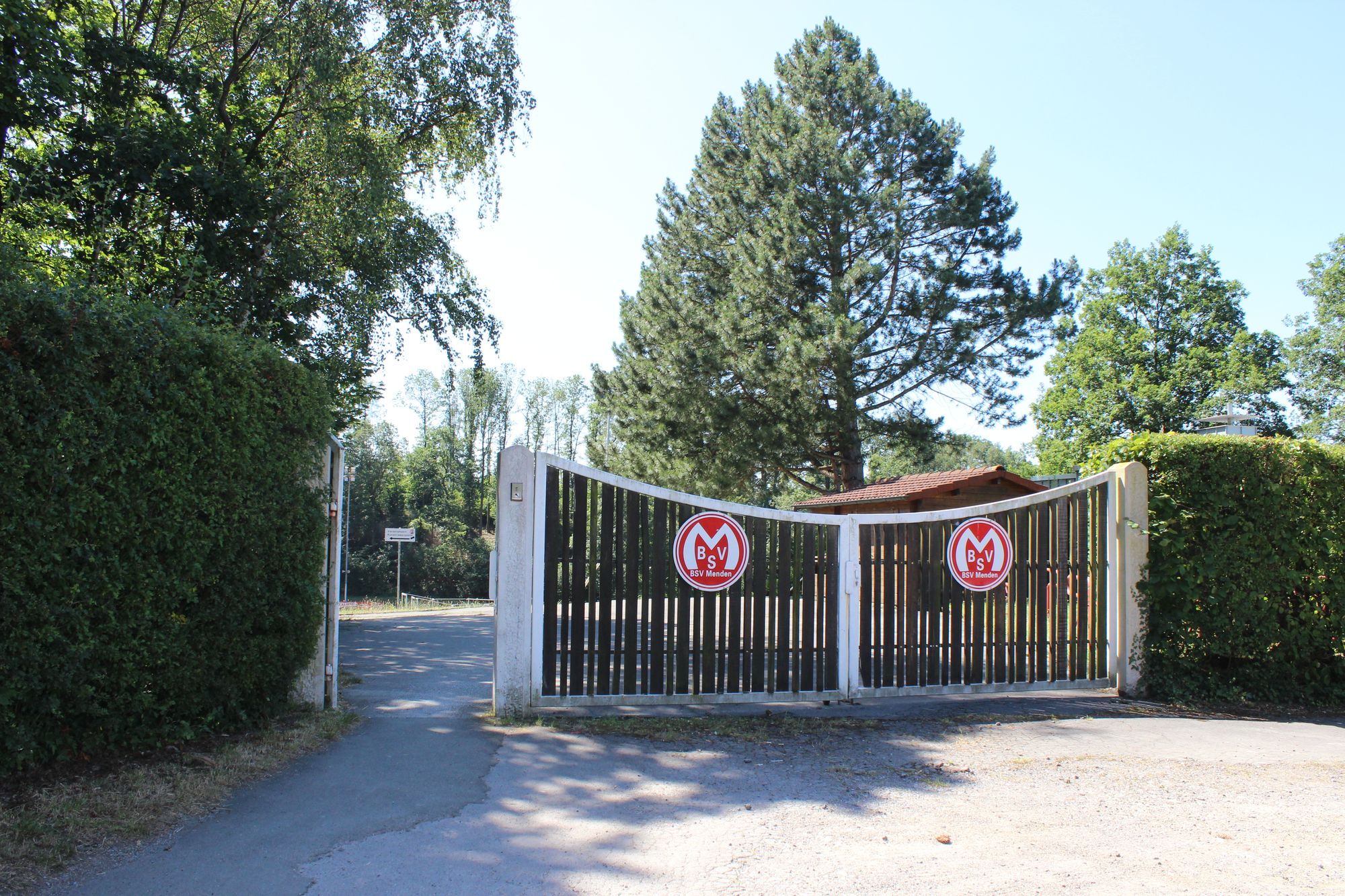 Das Bild zeigt ein Eingangstor mit zwei runden rot-weißen Wappen des BSV. Nebenan sind Hecken. Ein Tor ist offen, das große geschlossen.