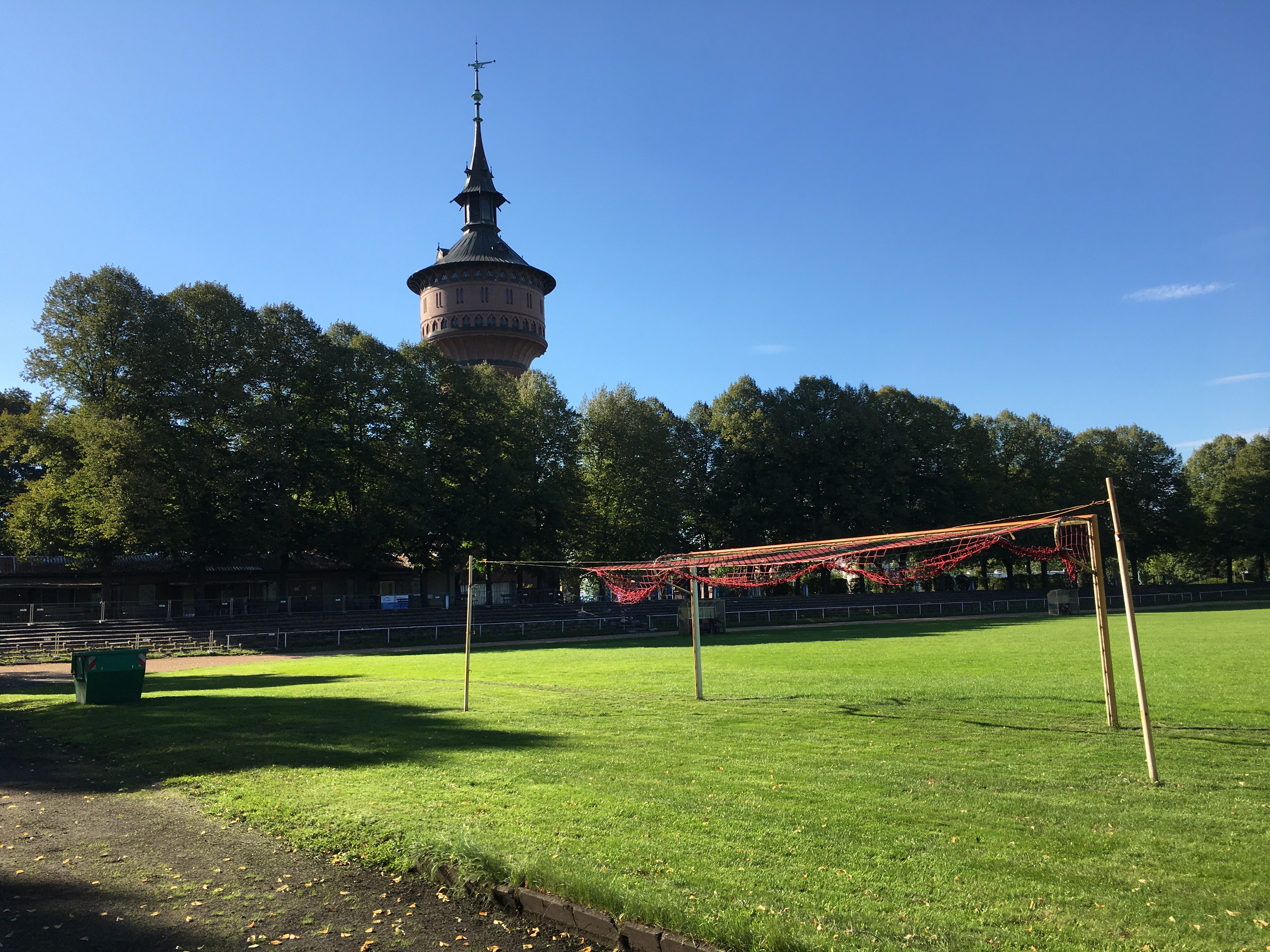 Das Bild zeigt einen Fußballplatz. Im Vordergrund ist ein Tor zu sehen, dessen Netz sehr zerschlissen ist. Im Hintergrund stehen Bäume und ein alter Wasserturm.