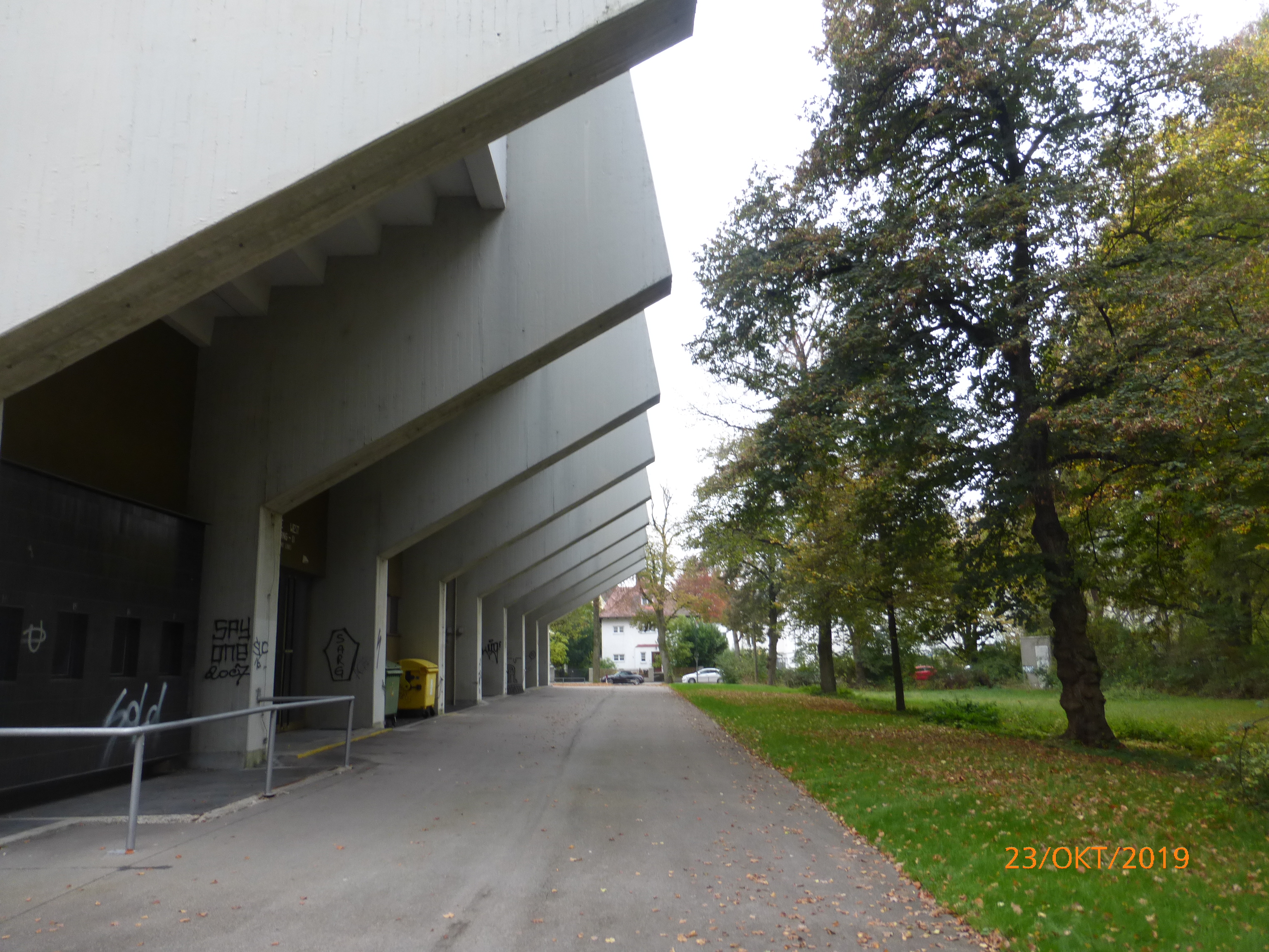 Das Bild zeigt eine Sporthalle, vor der ein Fußweg verläuft. Auf der rechten Seite befindet sich eine Wiese mit Bäumen.