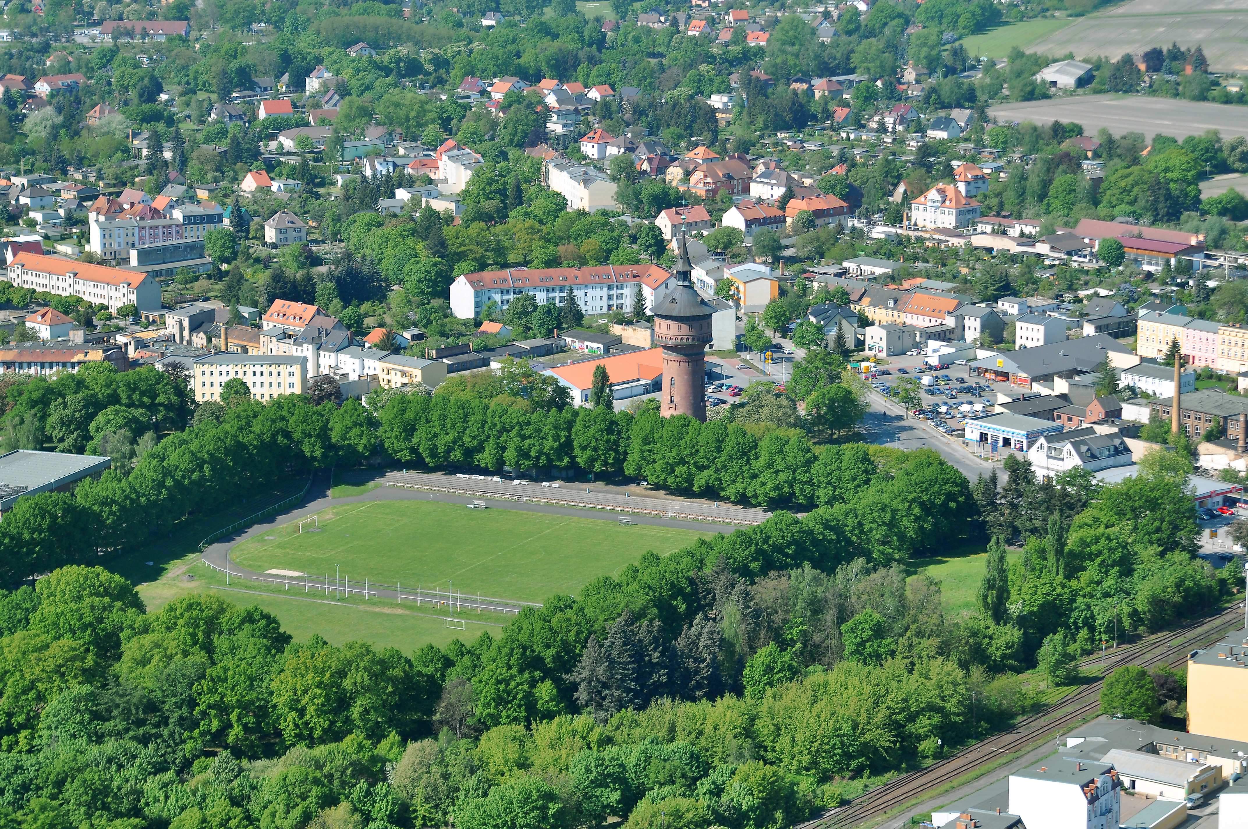 Die Luftaufnahme zeigt das Sportzentrum von oben. Rundherum ist der Sportplatz von Bäumen umgeben. Im Hintergrund sieht man eine Stadt.